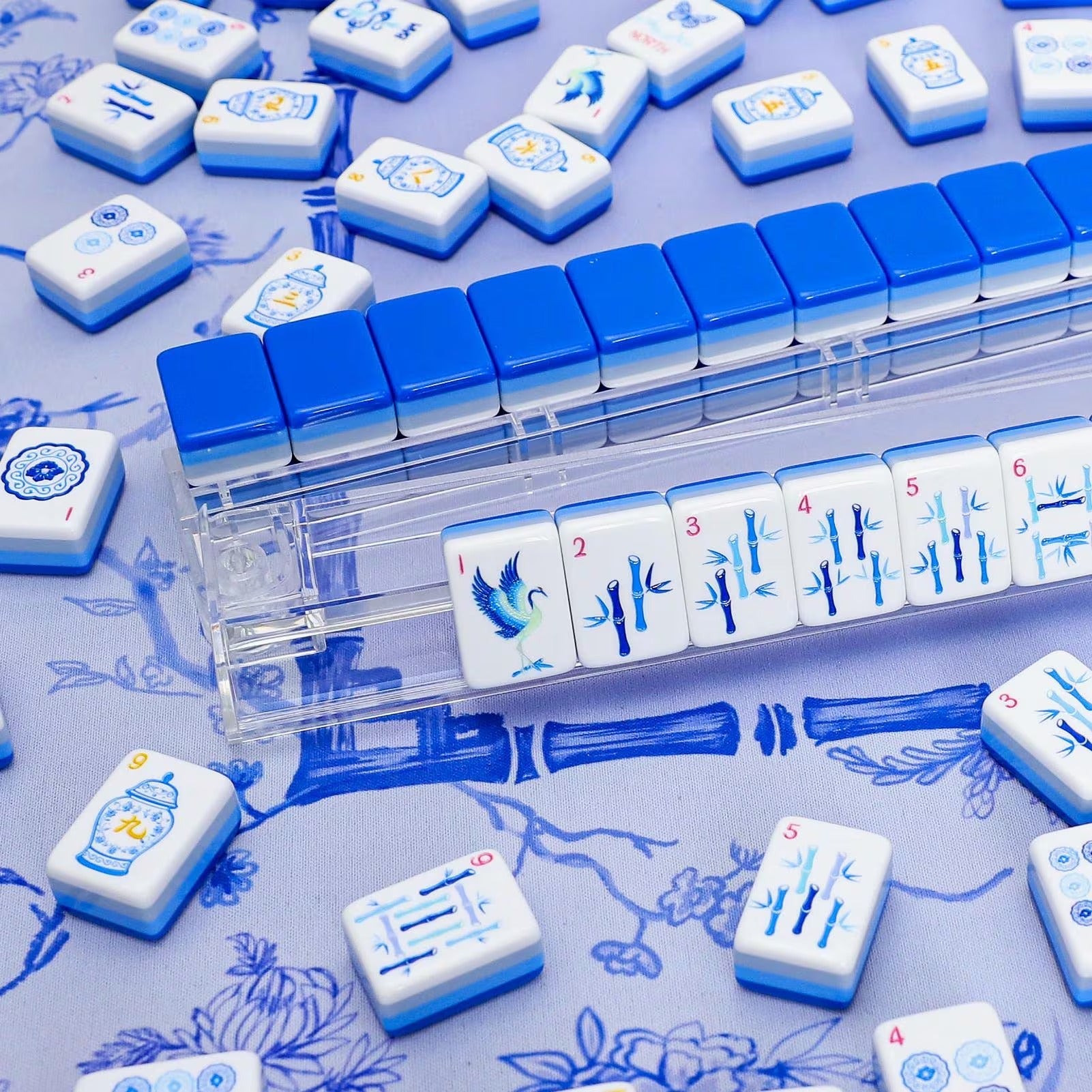 Classic blue and white porcelain style American Mahjong bamboo tiles lined up on a clear acrylic rack with a background of scattered gradient-stripe tiles.